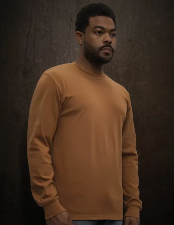 Portrait of a man with short curly hair and a beard, wearing a tan crewneck sweatshirt, standing against a dark textured wall.