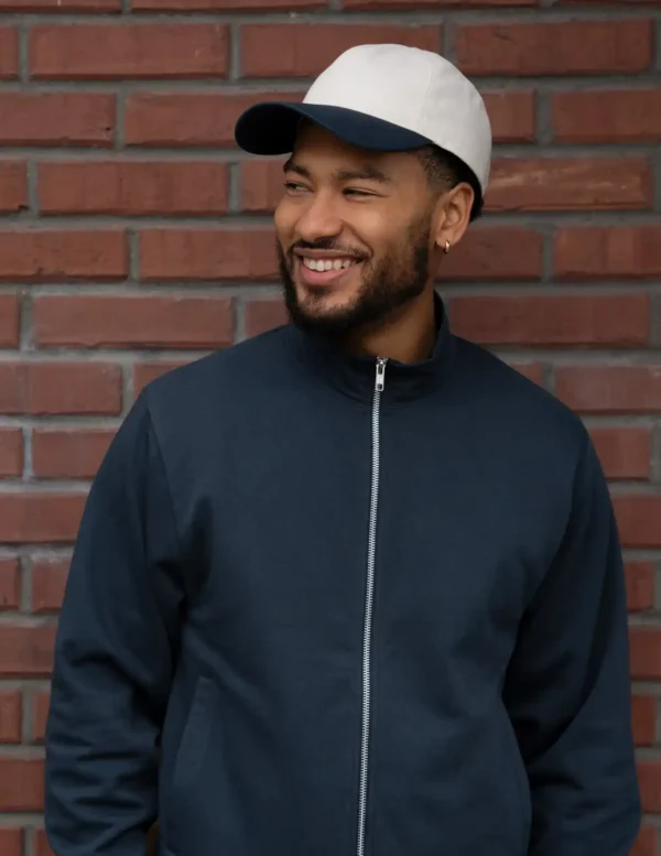 Smiling man in a dark zip-up jacket stands against a brick wall, wearing a white cap with a navy brim.